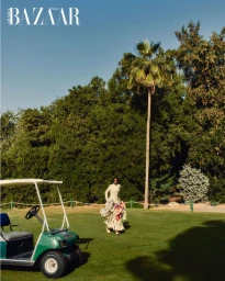 Model in a floral dress walking on a golf course near a green golf cart under a palm tree for Harper's Bazaar Arabia