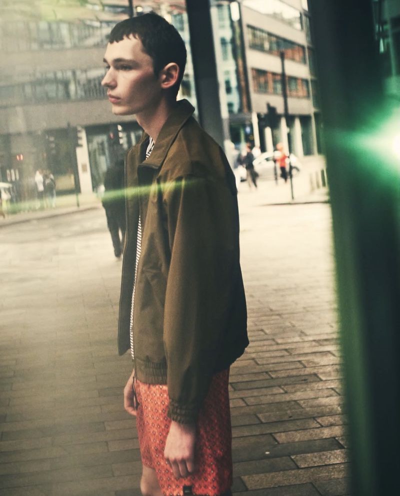 Profile view of a young man in a jacket and shorts, standing on a city street with blurred buildings and pedestrians in the background.