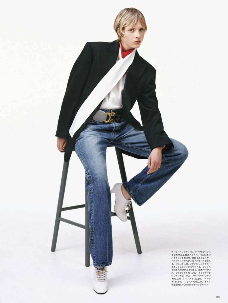 Model wearing a black blazer, white blouse, and blue jeans sitting on a stool against a plain white background.