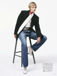 Model wearing a black blazer, white blouse, and blue jeans sitting on a stool against a plain white background.