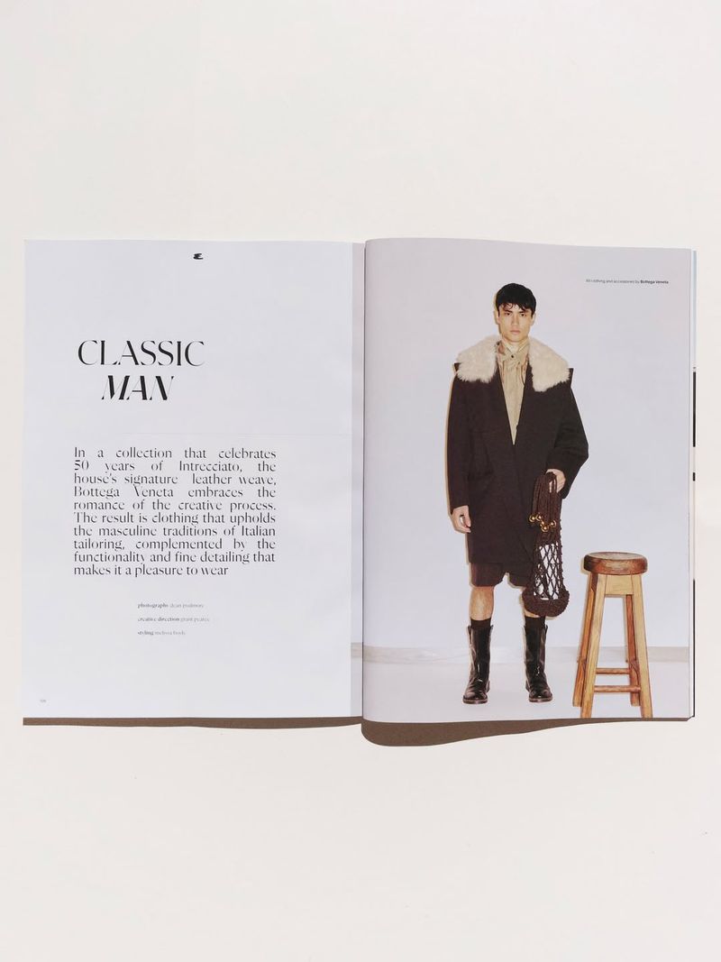 Magazine spread featuring a male model wearing a coat, shorts, and boots, posing next to a wooden stool against a white background.
