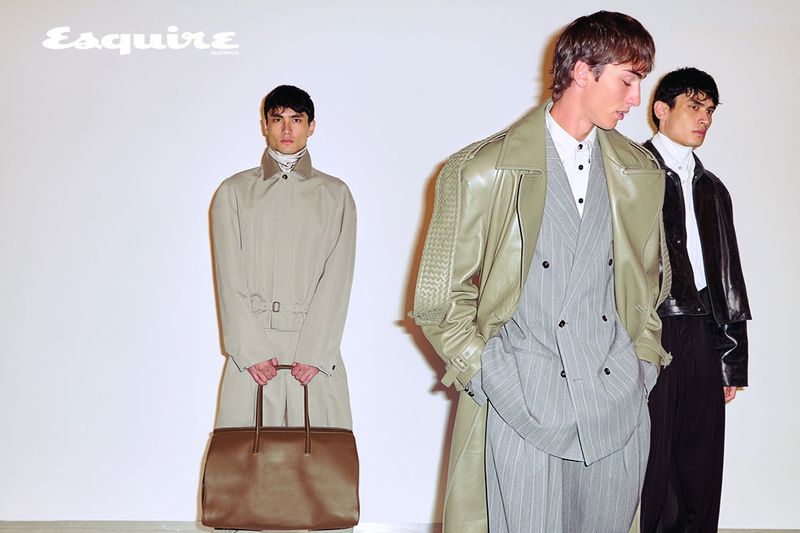 Three male models posing in designer menswear with a large brown leather bag against a clean white studio backdrop