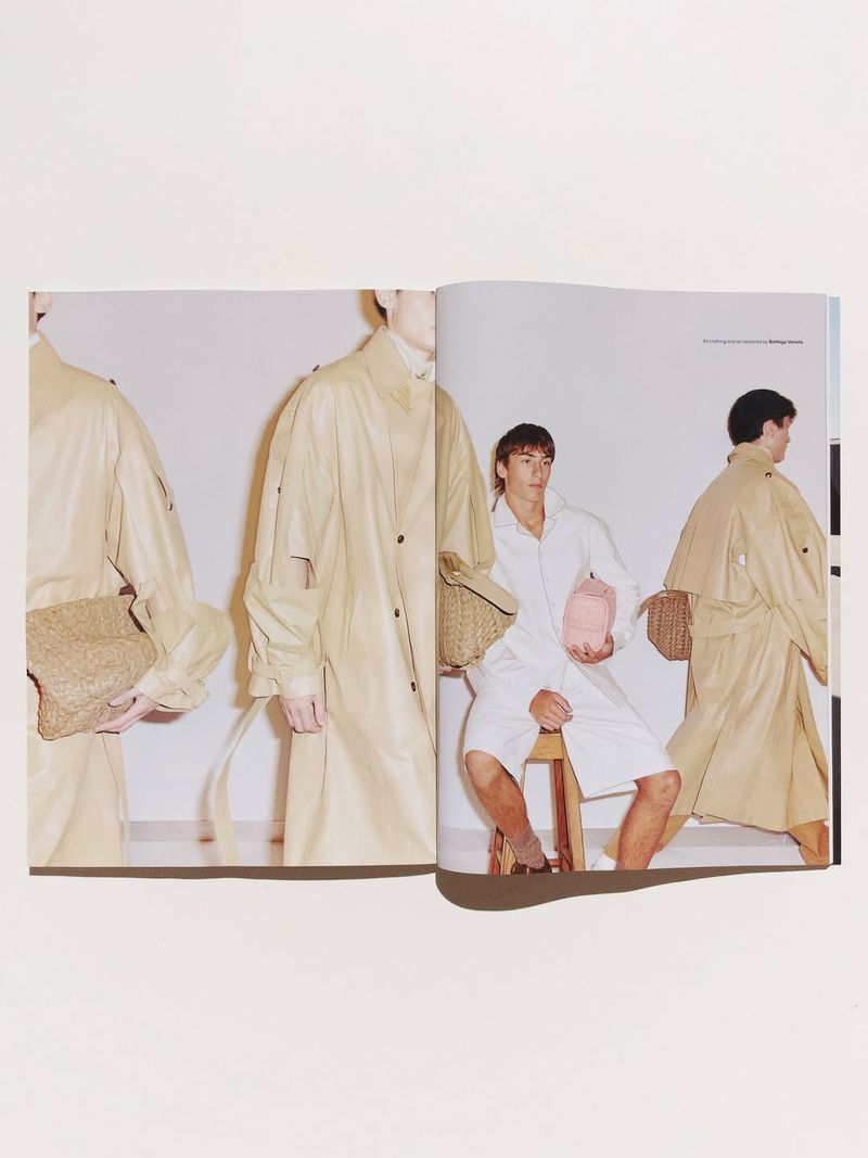 Magazine spread featuring a man seated on a stool wearing a white shirt and shorts alongside beige coats and woven bags.