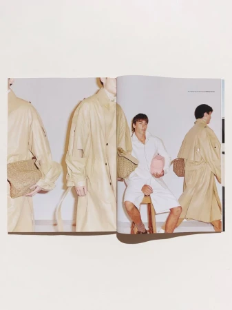 Magazine spread featuring a man seated on a stool wearing a white shirt and shorts alongside beige coats and woven bags.