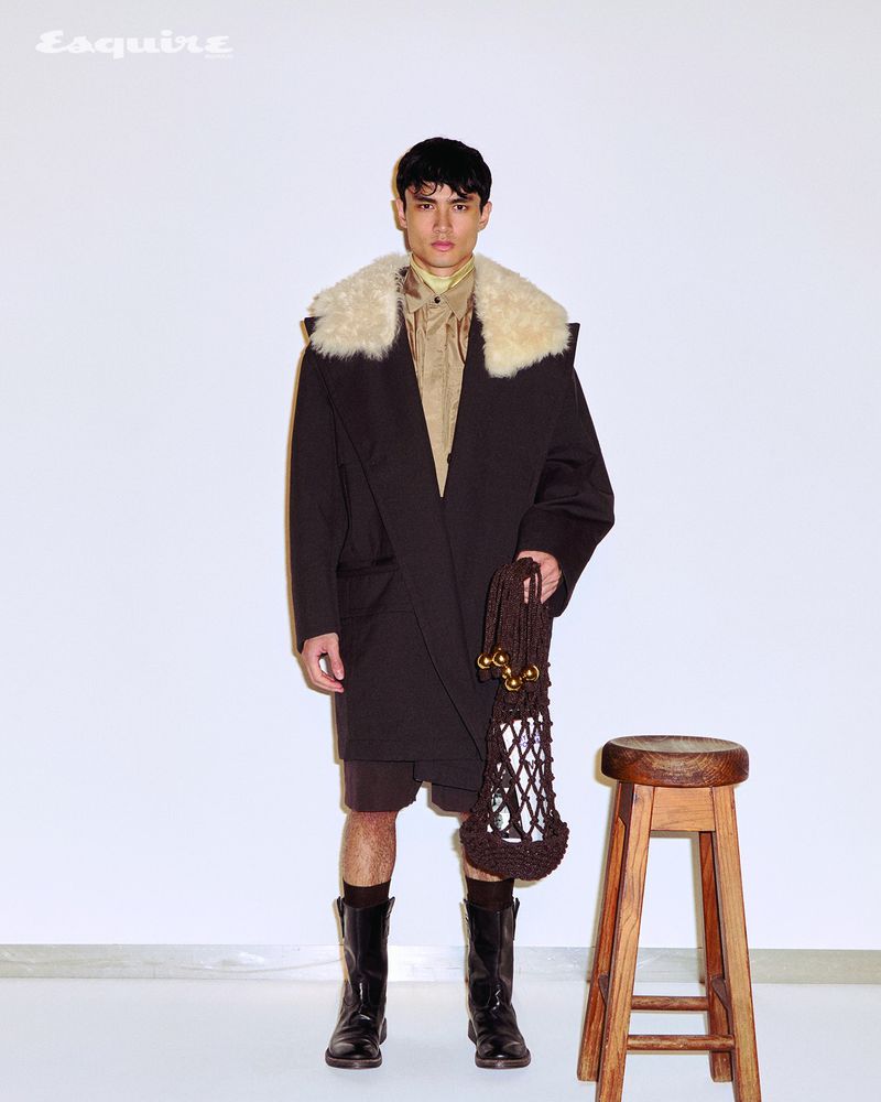 Man wearing a dark coat with a cream fur collar and holding a brown knit bag standing against a white seamless backdrop.
