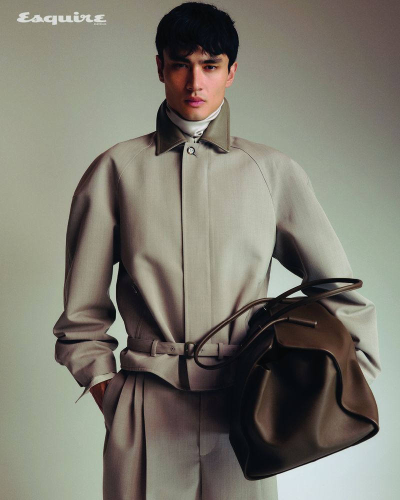 Man wearing a beige collared jacket and trousers holding a dark leather bag against a neutral studio background.