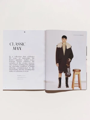 Double-page magazine spread featuring a male model in a dark coat with fur collar and tall boots standing beside a wooden stool