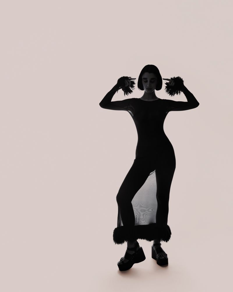 Model in dark sheer dress with fluffy cuffs pointing at head against a pale neutral background for Numero Netherlands.