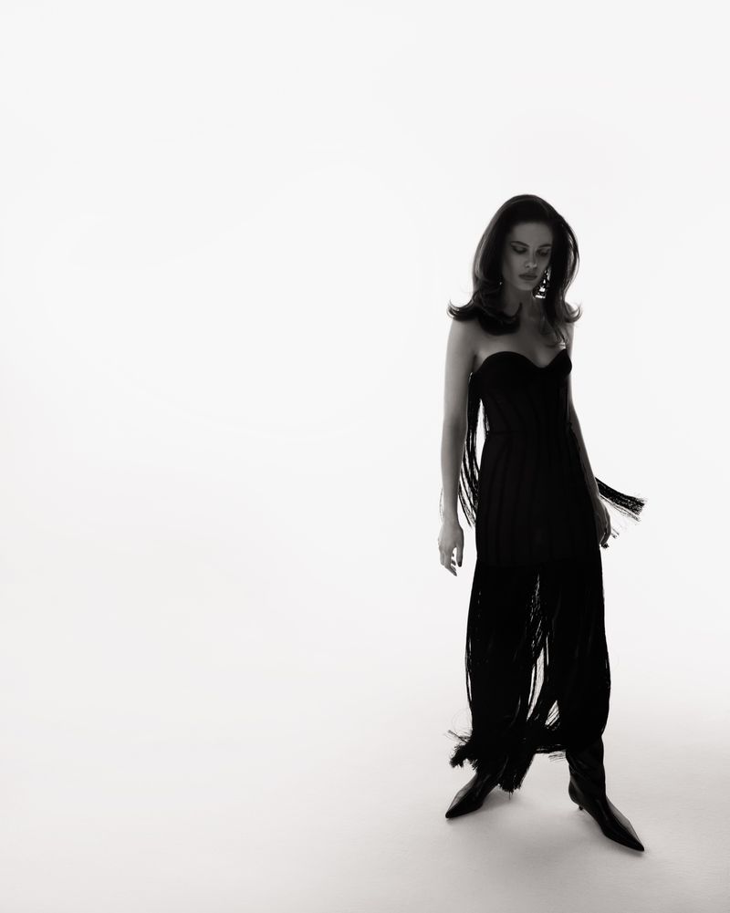 Model in a black fringe dress and boots standing against a bright white studio background.