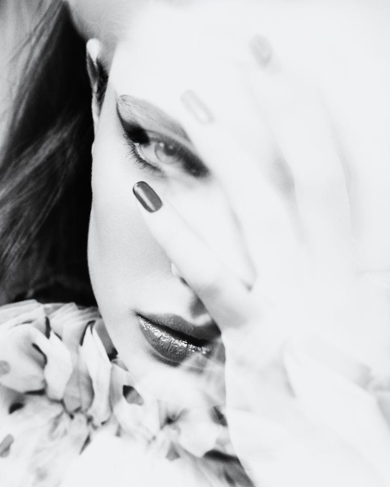 Black and white portrait of a woman with red lipstick covering half of her face with her hand, wearing a floral-patterned shirt.