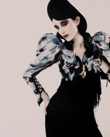 Model wearing a black dress with ruffled feather sleeves and a tall hat against a light background
