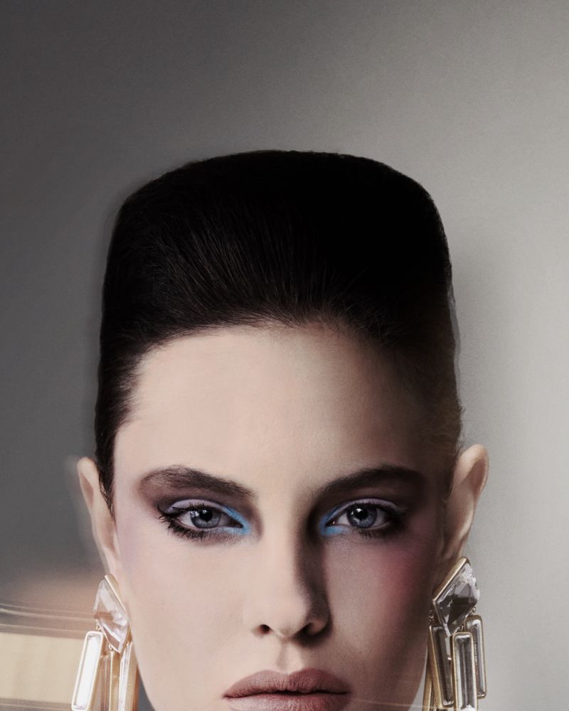 Close-up portrait of a model with voluminous dark hair, blue eyeshadow, and large crystal earrings against a neutral grey background.