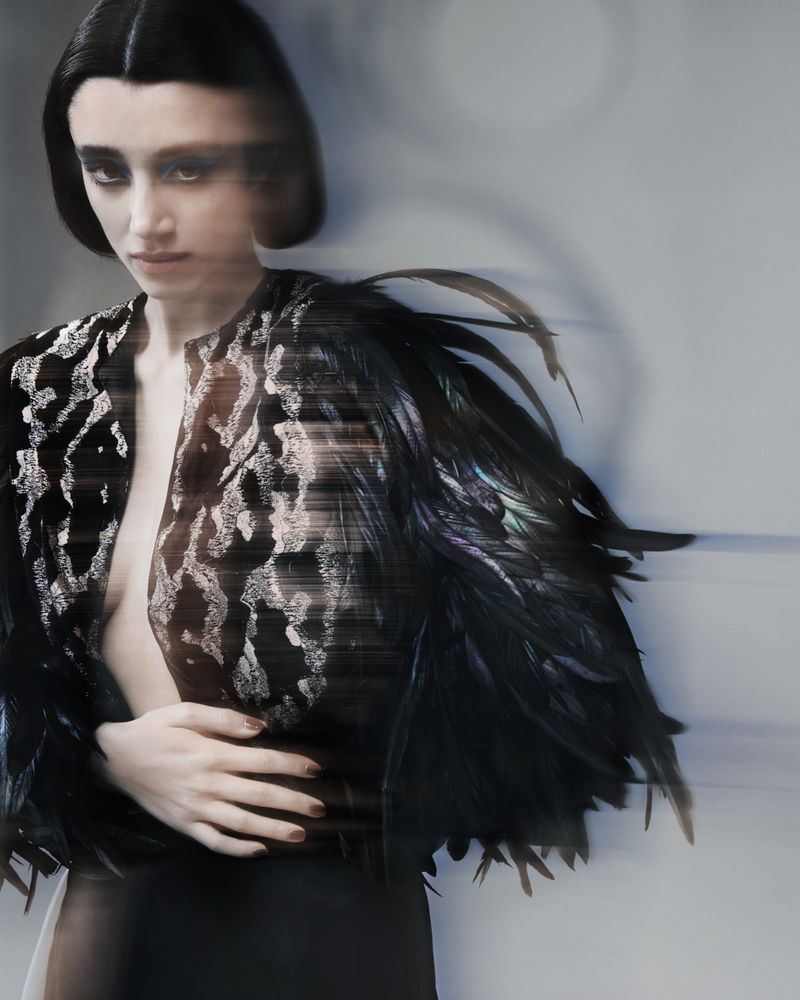 Model with motion-blurred dark hair wearing a black feather-adorned patterned top against a plain light grey background