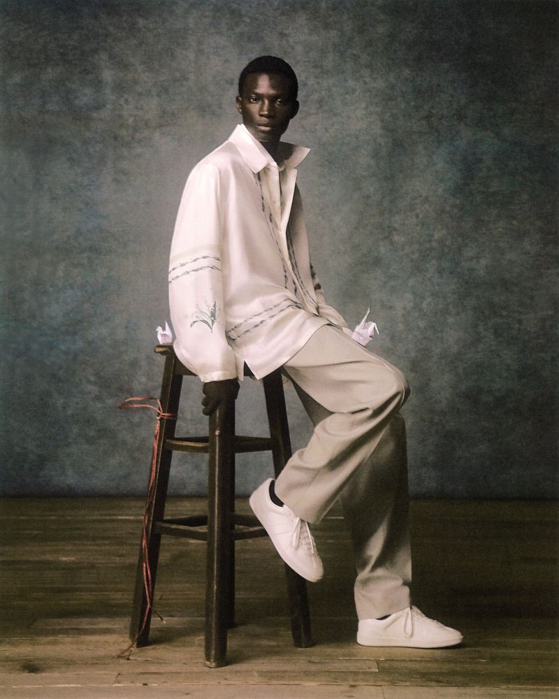 Studio shot of a dark-skinned male fashion model wearing a white button-down shirt and khaki pants, sitting on a stool, posing.