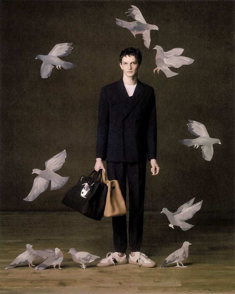 Elegant male model in a black suit surrounded by doves in a dark studio setting, holding black and tan tote bags, lifestyle fashion editorial.