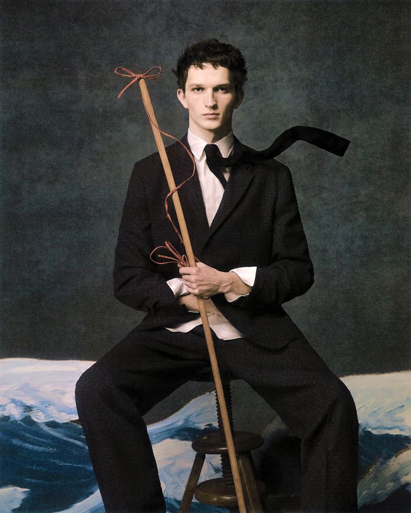 A young adult male in a dark suit sits holding a stick with ties in front of a cloudy background with painted waves.