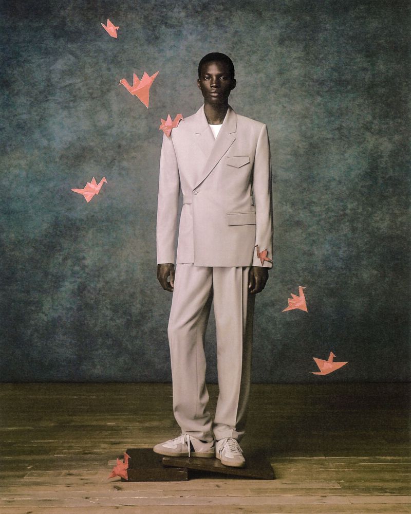Man stands on platform in suit with paper origami birds in studio against textured teal backdrop, in a fashion forward portrait.