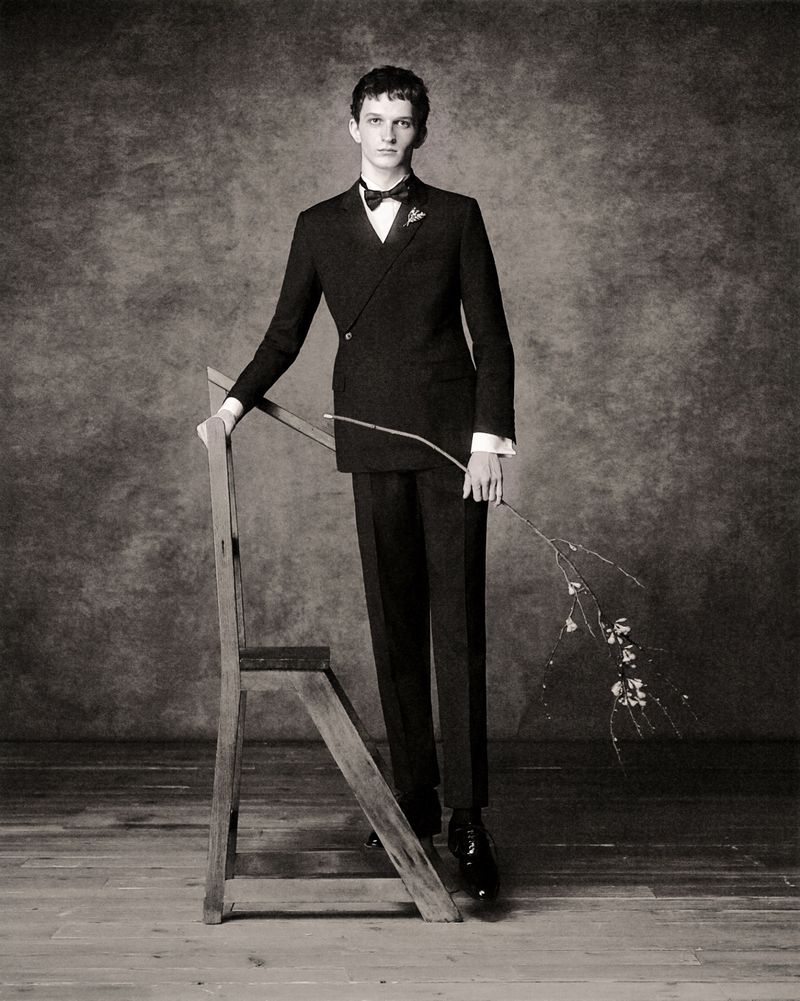 Elegant studio portrait of a tall, slender man in a black tuxedo, leaning on a vintage wooden ladder while holding a delicate branch.