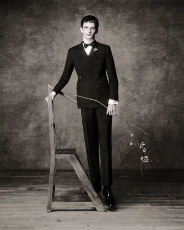 Elegant studio portrait of a tall, slender man in a black tuxedo, leaning on a vintage wooden ladder while holding a delicate branch.