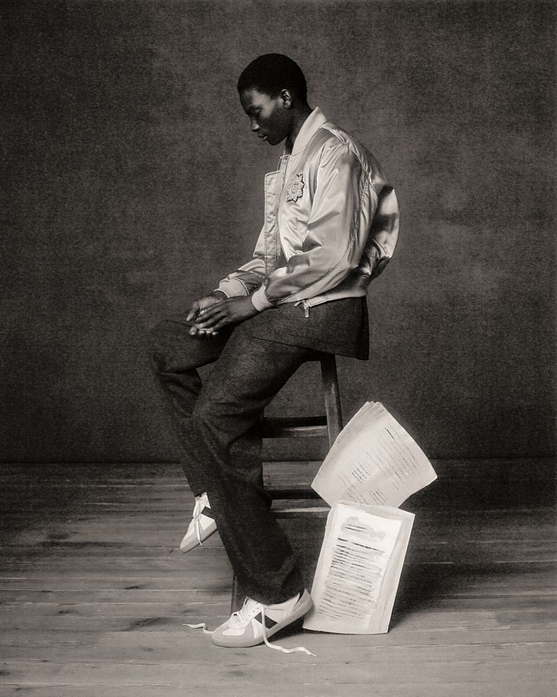 Black and white studio portrait of an African American male model in a bomber jacket sitting on a wooden stool with scattered papers.