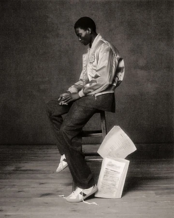 Black and white studio portrait of an African American male model in a bomber jacket sitting on a wooden stool with scattered papers.