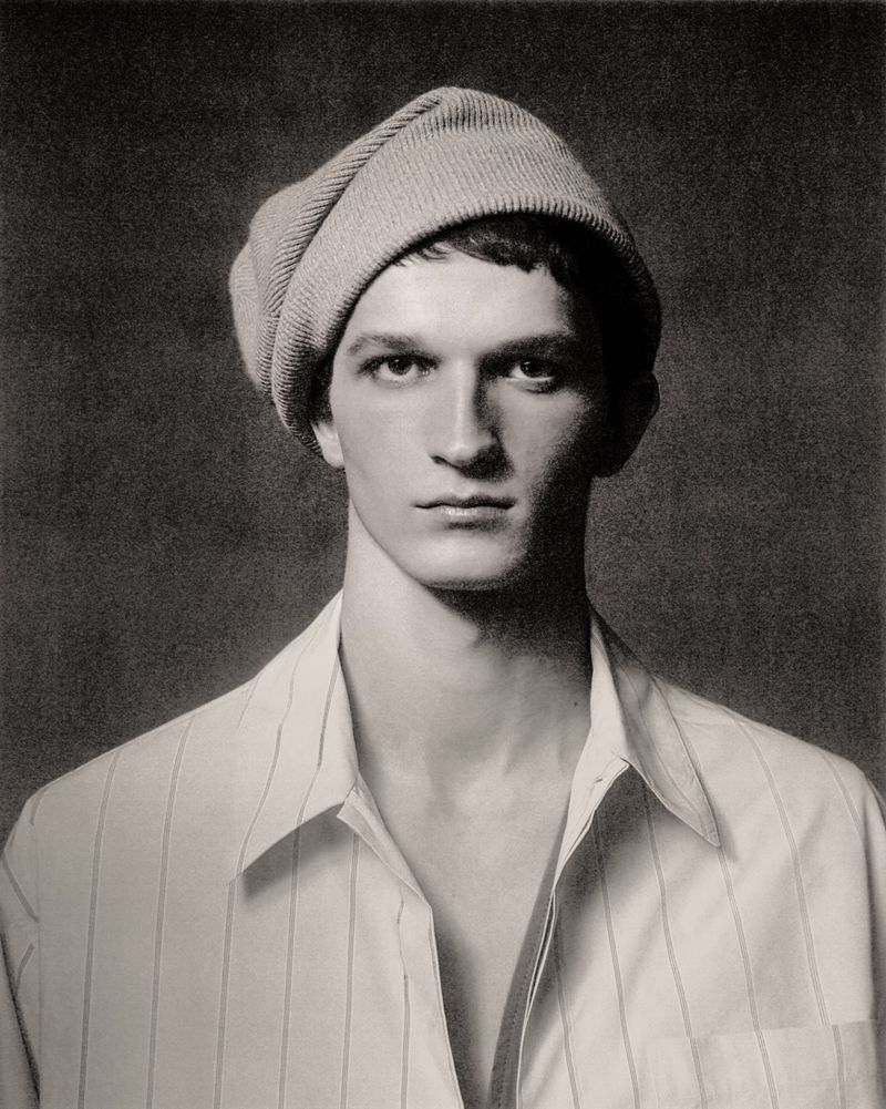 Portrait of a young adult male wearing a beanie and a white striped shirt, set against a textured, dark grey background.
