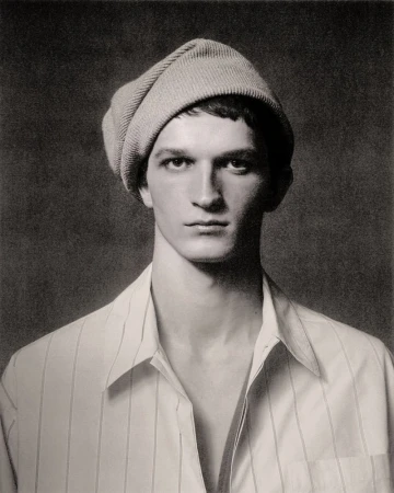 Portrait of a young adult male wearing a beanie and a white striped shirt, set against a textured, dark grey background.