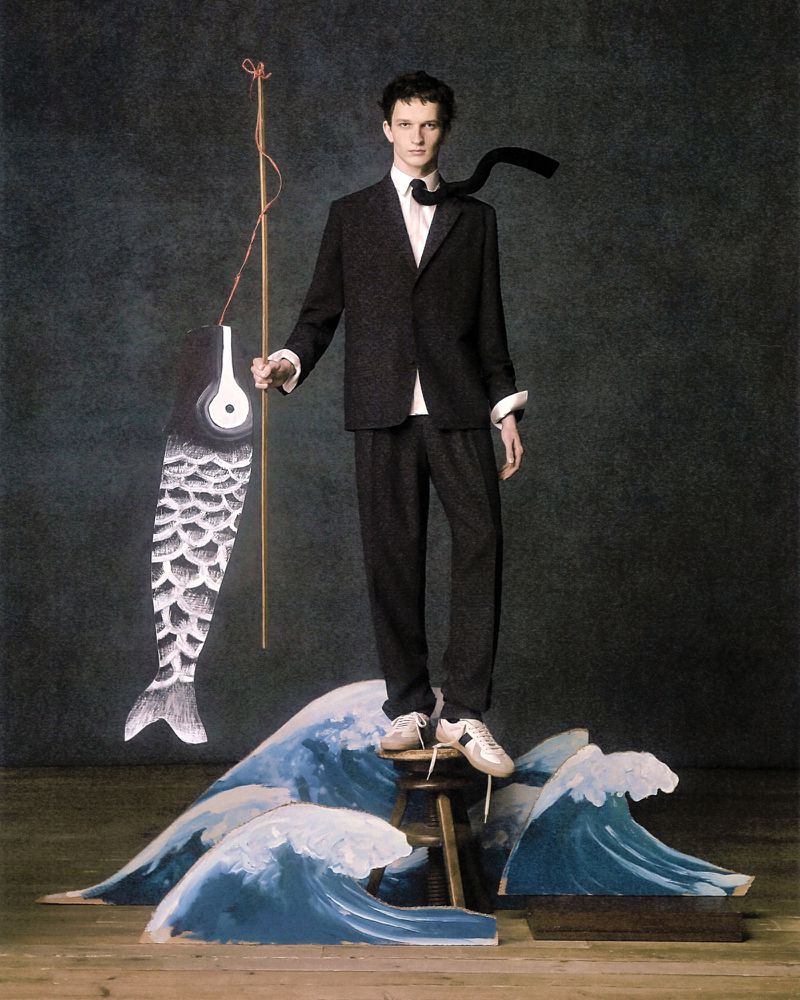 A slender adult male model poses on a wooden stool in a fashion editorial image, wearing a dark suit and tie, and holding a paper fish.