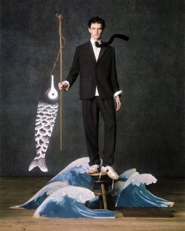 A slender adult male model poses on a wooden stool in a fashion editorial image, wearing a dark suit and tie, and holding a paper fish.