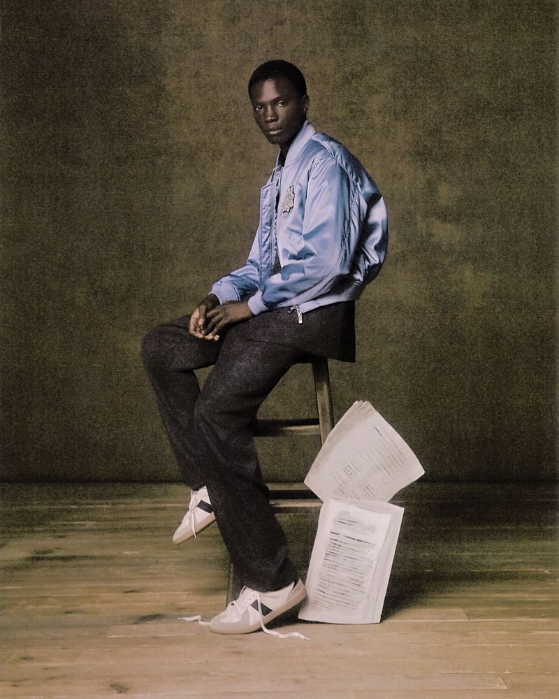A young, dark-skinned adult male poses indoors, seated on a wooden stool in a light blue jacket and dark pants with off-white sneakers, documents below.
