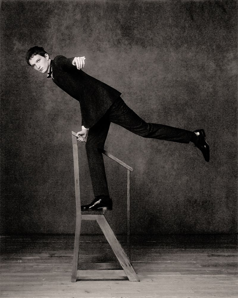 Young man in a suit posing on a wooden step stool in a sepia-toned studio setting, balancing with one leg extended. Fashion photography.