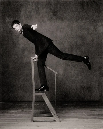 Young man in a suit posing on a wooden step stool in a sepia-toned studio setting, balancing with one leg extended. Fashion photography.