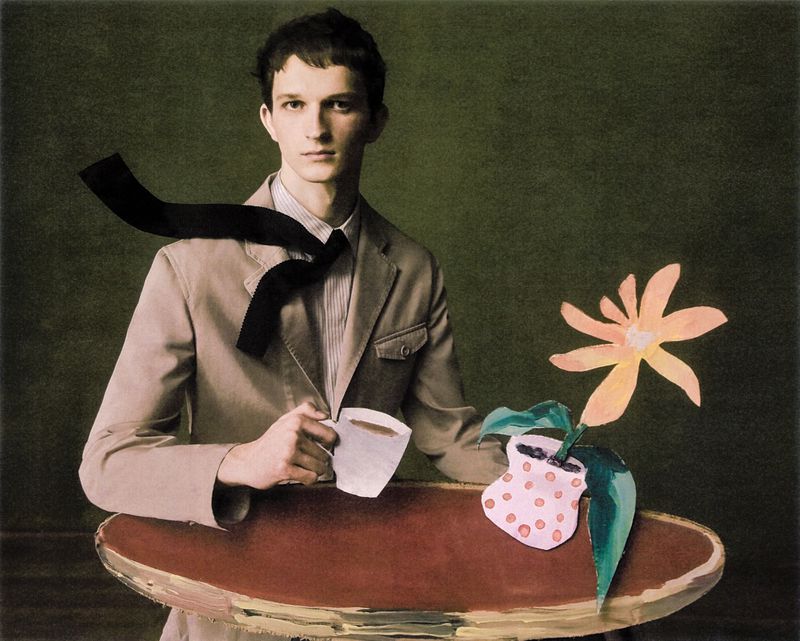 Man in a suit holding a coffee cup, next to a drawing of a flower in a pot, against a green background in an indoor setting.