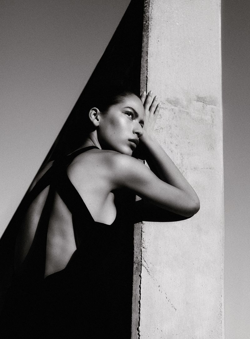 Model in black dress leaning against a concrete pillar outdoors in monochrome editorial fashion portrait