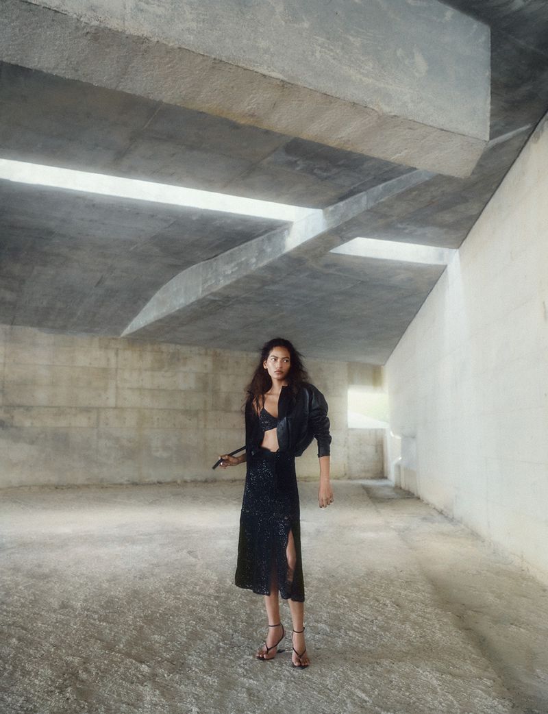Model wearing dark jacket and sequin skirt standing in a concrete industrial architectural space with overhead beams.