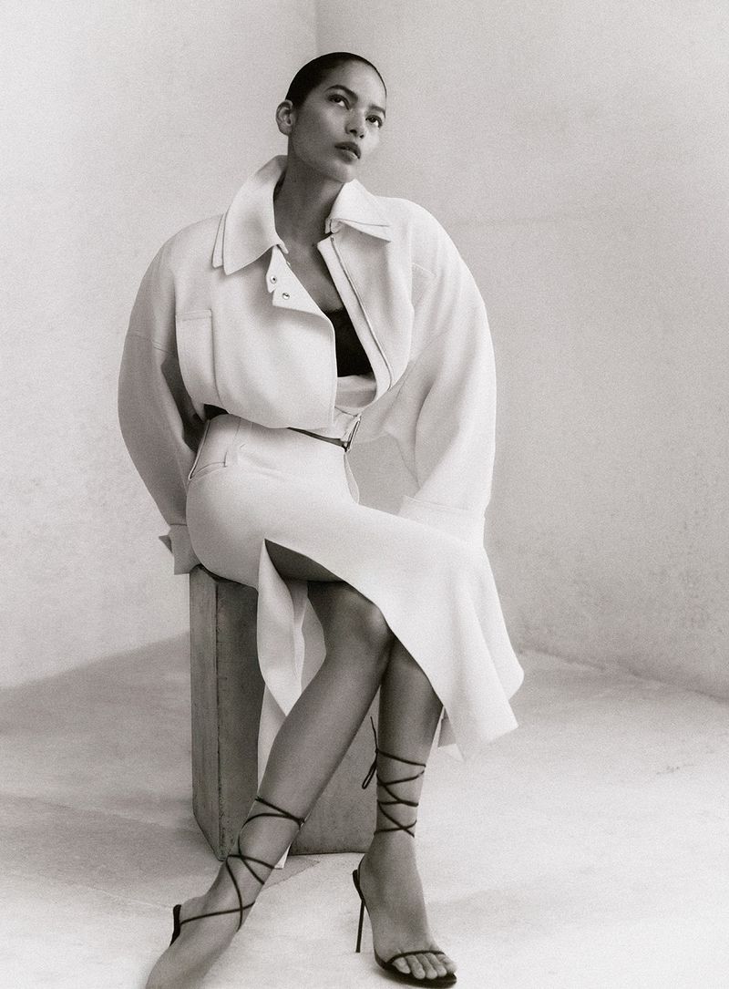 Monochrome fashion editorial portrait of a model seated on a block wearing a light-coloured collared jacket and skirt with lace-up heels