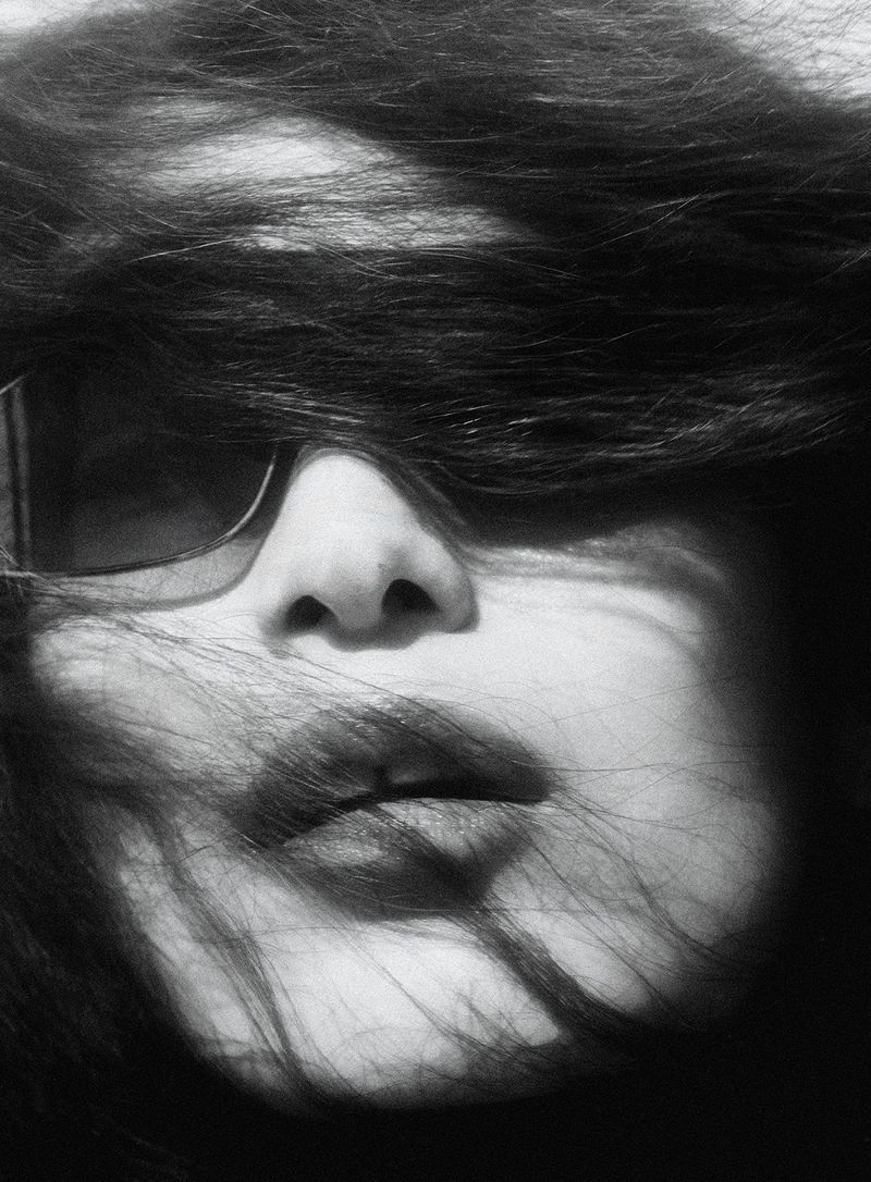 Close up portrait of a model with dark hair blowing across her face and eyes partially obscured by sunglasses in a high contrast black and white photo