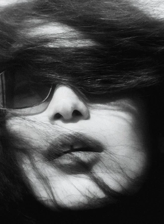 Close up portrait of a model with dark hair blowing across her face and eyes partially obscured by sunglasses in a high contrast black and white photo