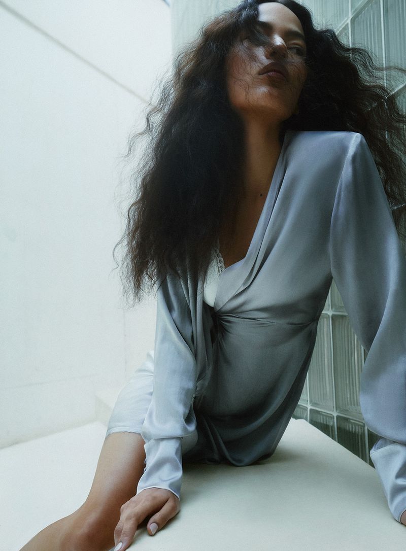 Model with long dark wavy hair wearing a grey satin dress seated against an architectural white wall for Vanity Fair Italia