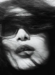 Close up portrait of a model with dark hair blowing across her face and eyes partially obscured by sunglasses in a high contrast black and white photo