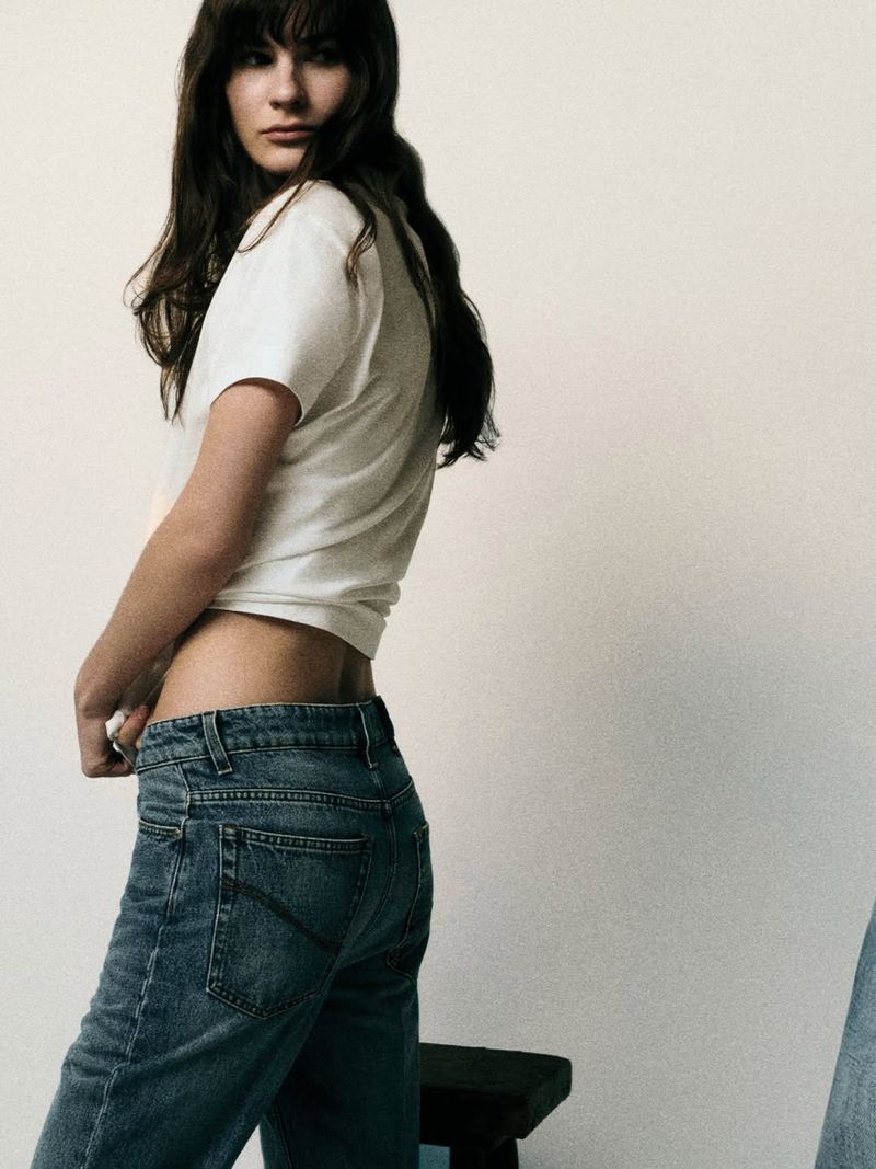 Model with long dark hair wearing a white t-shirt and blue denim jeans against a plain neutral background