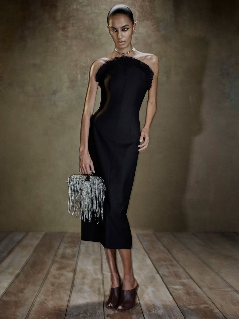 Model in black strapless dress holding a fringed bag standing on wooden floor