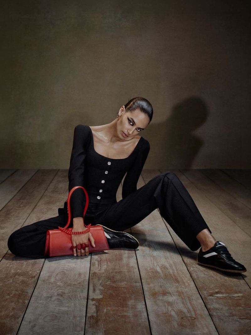 Model seated on a wooden floor wearing black clothing and holding a red handbag for Alexander McQueen