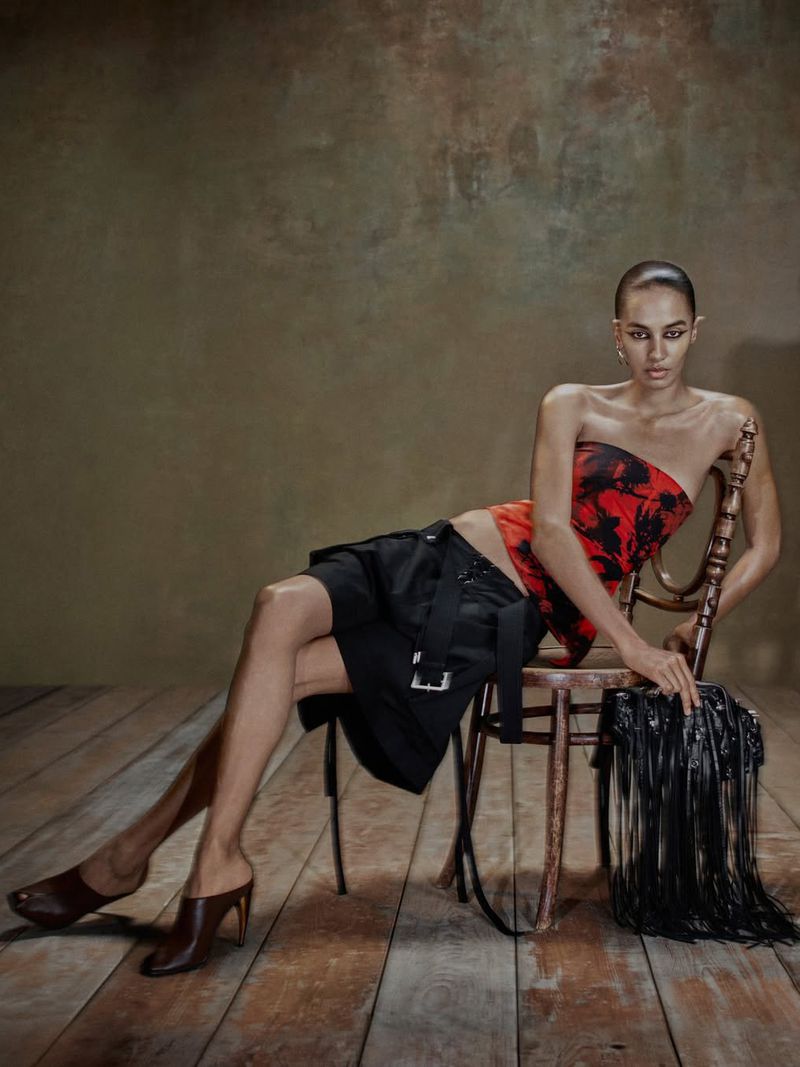 Model seated on a wooden chair wearing a red printed strapless top and black shorts against a textured backdrop