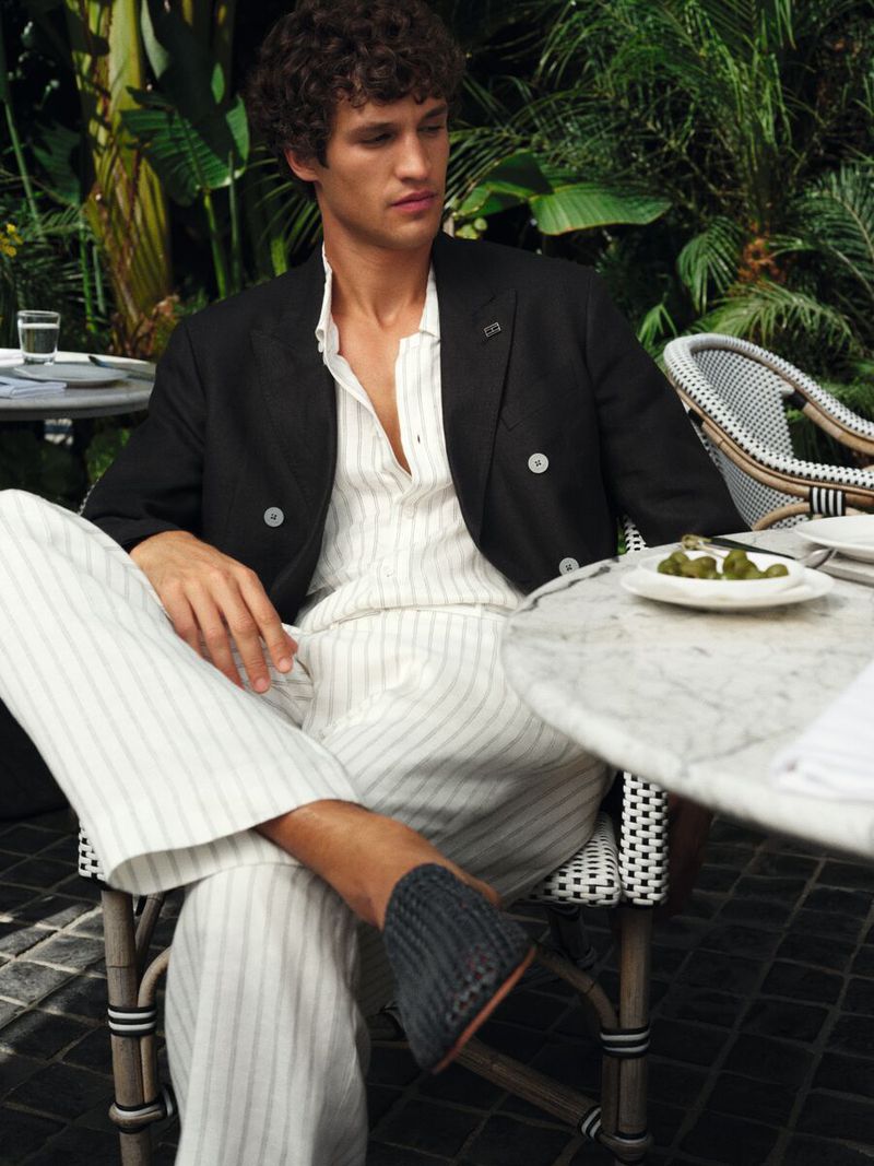 Model seated outdoors at a cafe wearing a black blazer and white linen striped trousers for Tommy Hilfiger.