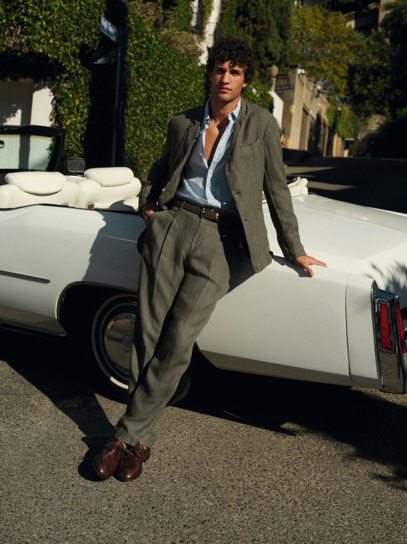 Male model in an olive green suit leaning against a vintage white convertible car on a sunny street.