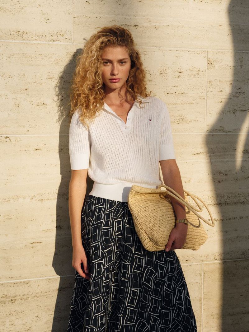 Model with long curly blonde hair wearing a white polo shirt and patterned skirt holding a woven straw tote bag against a stone wall