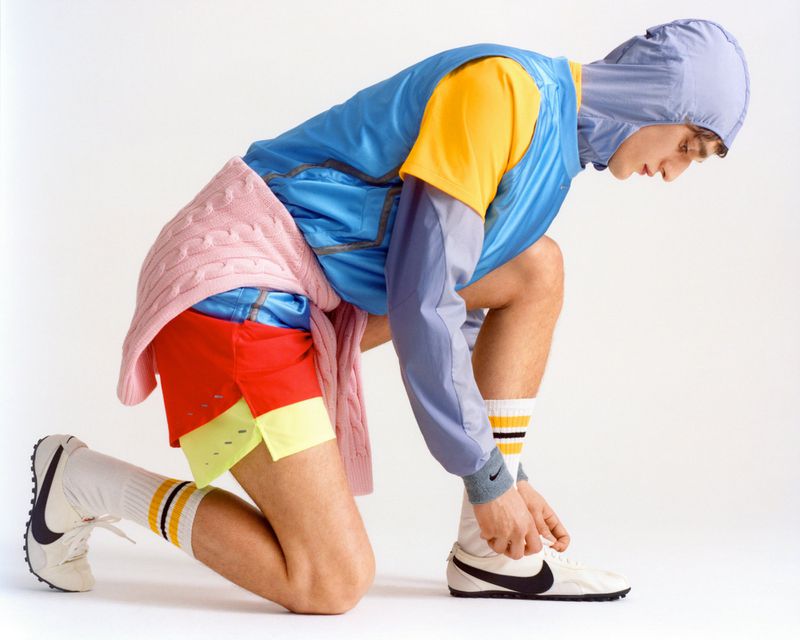 Profile of a male model kneeling on a white background, wearing a blue hooded top, yellow shirt, red shorts, and white sneakers.
