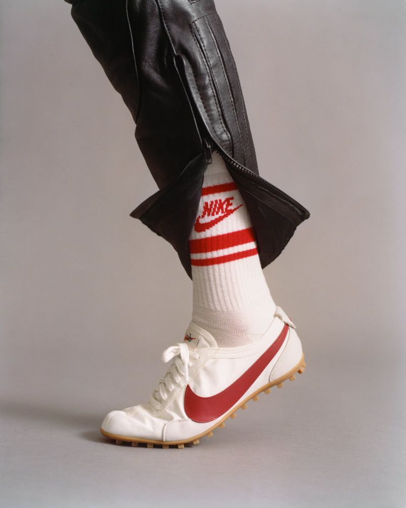 Close-up shot of a white sneaker with red swoosh, striped Nike socks, and black leather trousers against a grey seamless background