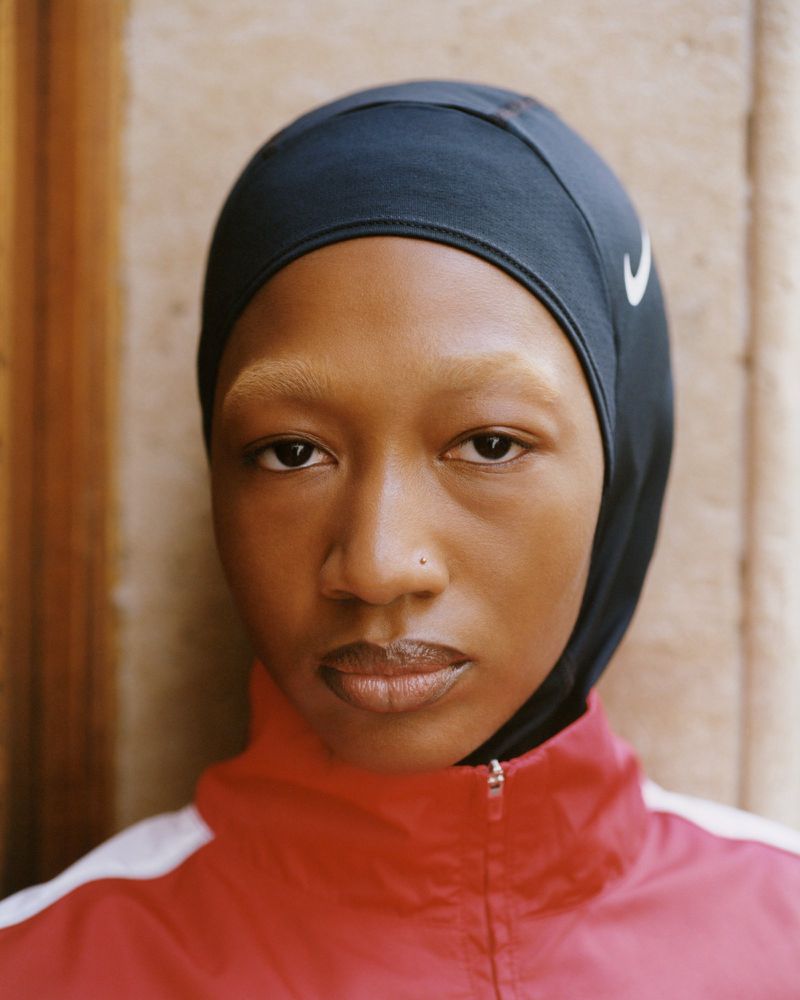 Close-up portrait of a Black woman wearing a black athletic head covering with a white swoosh logo and a red jacket.